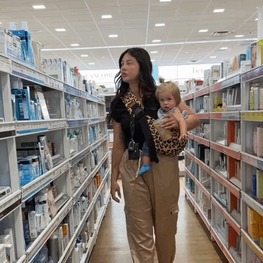 Mother browsing a pharmacy aisle while carrying her baby in the Loravi Sling