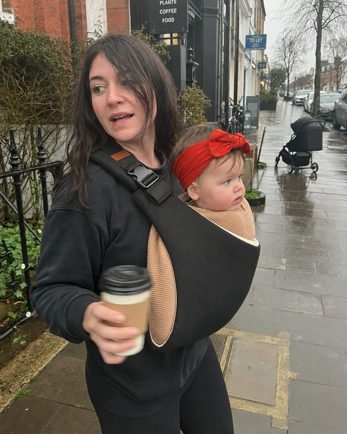 Mother holding a coffee while carrying her baby in the Loravi Sling on a rainy street